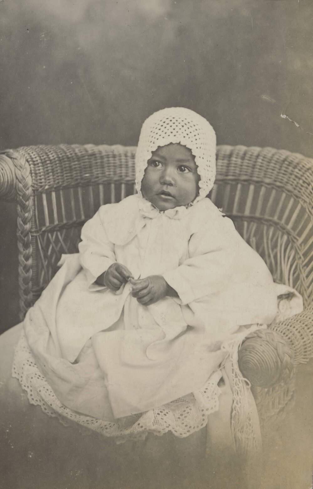 Baby wearing bonnet and seated in a cane chair