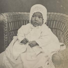 Baby wearing bonnet and seated in a cane chair